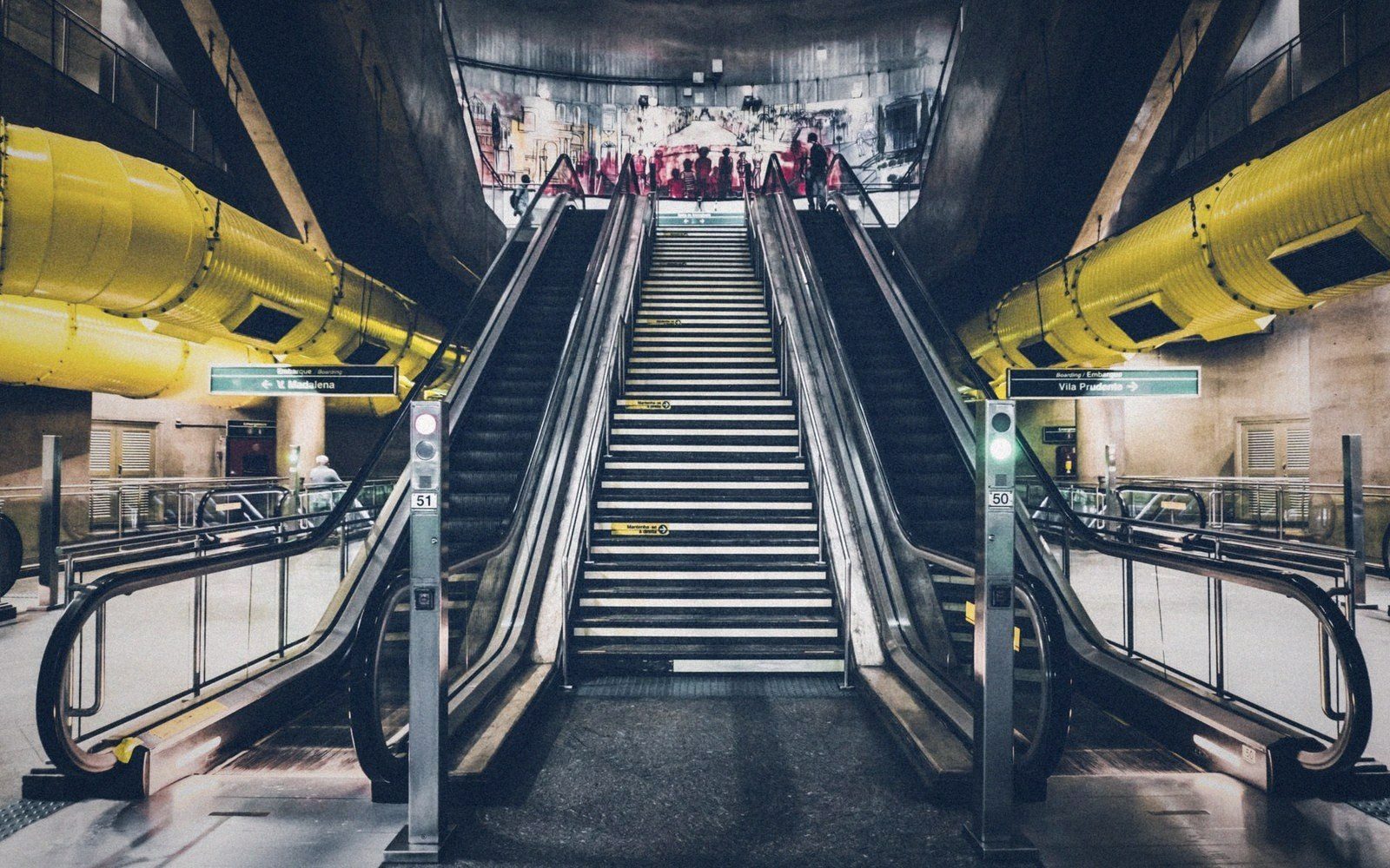 Escalators flank a central staircase in a subway station with yellow overhead ducts