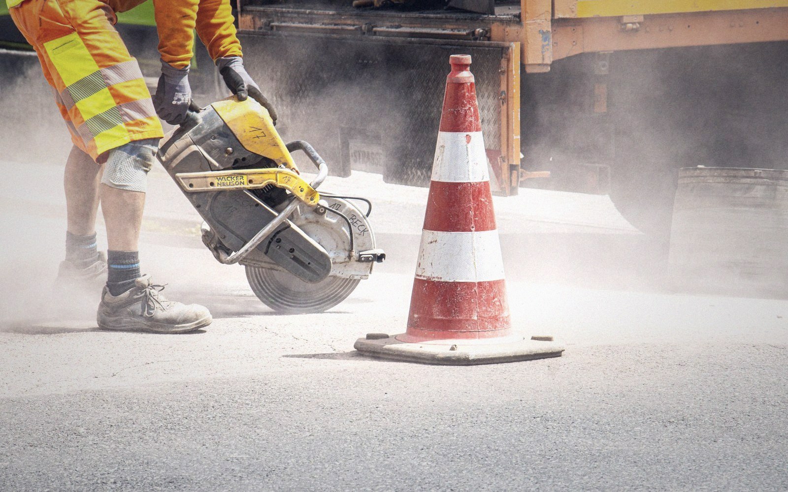 Worker cuts pavement with a gas-powered saw beside a traffic cone amid dust
