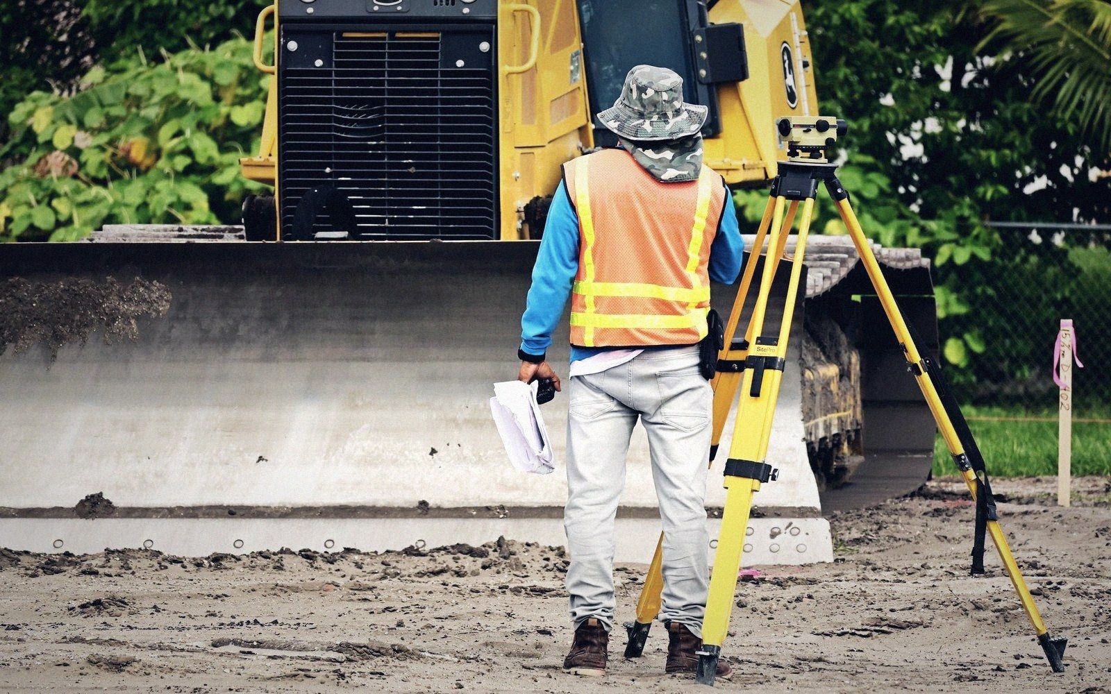 Surveyor in a safety vest stands beside a tripod near a bulldozer at a construction site